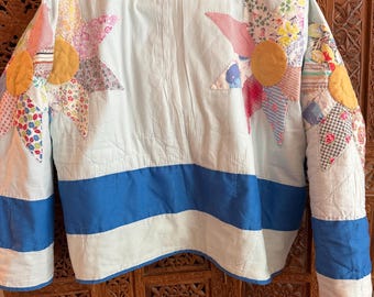 Puede incluir: Una sudadera con capucha de color azul claro confeccionada en tela acolchada. La prenda presenta grandes aplicaciones en forma de estrella con motivos florales y estampados en el pecho y las mangas, con centros circulares amarillos y rayas azules.