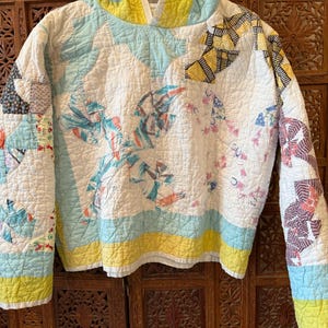 May include: A quilted jacket with a unique patchwork design. The jacket features a white base with patches of blue, yellow, and patterned fabrics. The collar and cuffs are trimmed in yellow. The jacket is hanging on a black hanger.