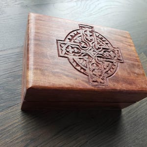 May include: A rectangular wooden box with a carved Celtic cross design on the lid. The box is made of dark brown wood and has a smooth, polished finish. The cross is intricately carved with detailed patterns.