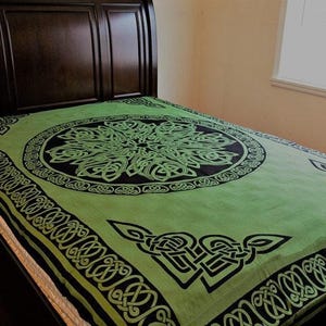 May include: A green bedspread with a black Celtic knot design. The bedspread features a large central knot pattern and border designs. The bedspread is on a bed with a dark brown headboard.