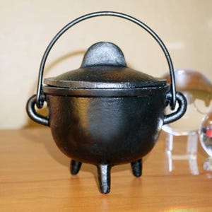 May include: A small, black cast iron cauldron with a matching lid and a curved handle. The cauldron has three short legs and is designed to sit on a flat surface. The cauldron is likely used for decorative purposes or for small-scale cooking.