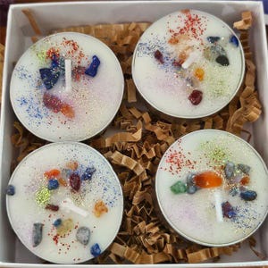 May include: Four white tea light candles with colorful gemstones and glitter. Each candle has a small wick and is set in a silver metal container. The candles are arranged in a white box with brown paper filler.
