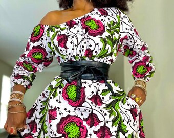 May include: A white dress with a pink and green floral print. The dress has a cinched waist with a black leather belt. The dress has a slightly off-the-shoulder neckline and long sleeves.