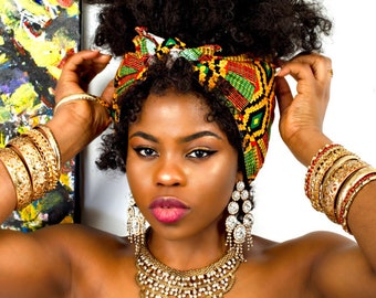 Puede incluir: Una mujer con cabello rizado marrón oscuro que lleva una diadema colorida, un collar de oro con piedras blancas y pulseras de oro. Lleva una blusa con estampado colorido.