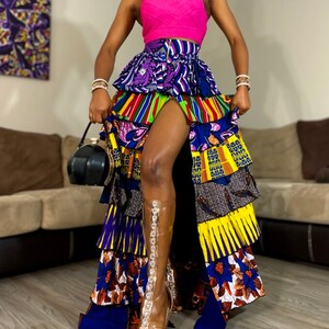 May include: A long, multi-colored skirt with a high-waisted fit and a ruffled design. The skirt is made of a patterned fabric with a variety of colors, including blue, yellow, green, and red. The skirt has a slit up the front and is paired with a pink tank top and clear heeled sandals.