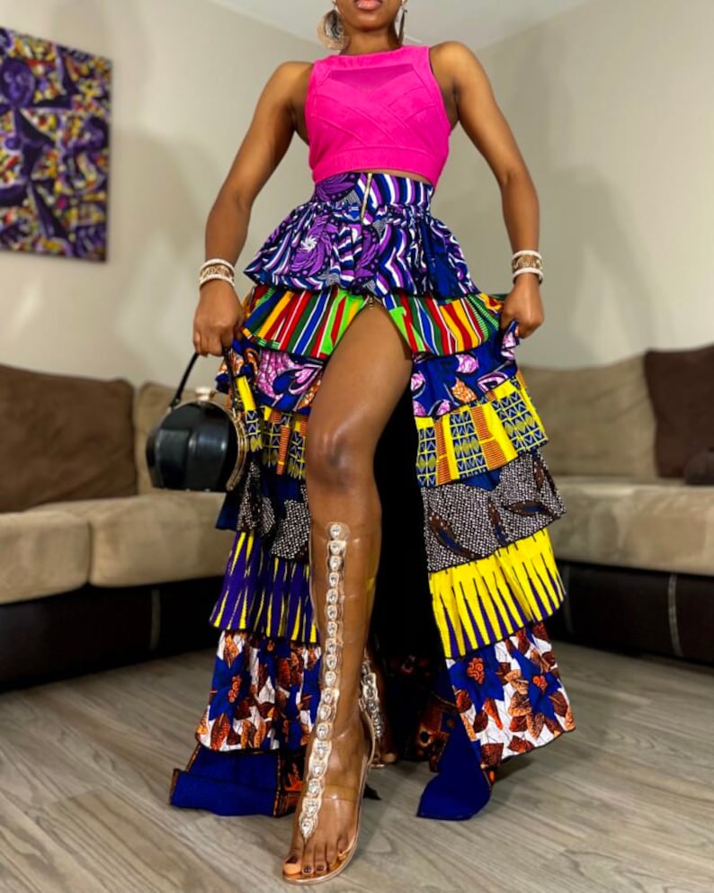 May include: A long, multi-colored skirt with a high-waisted fit and a ruffled design. The skirt is made of a patterned fabric with a variety of colors, including blue, yellow, green, and red. The skirt has a slit up the front and is paired with a pink tank top and clear heeled sandals.