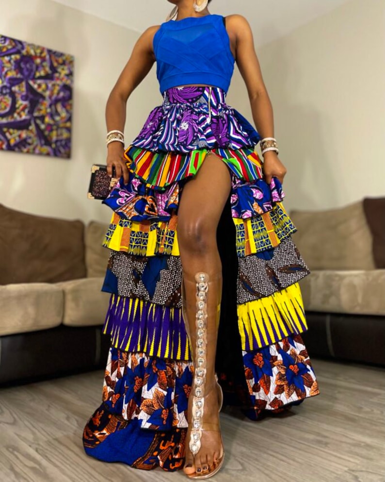 May include: A multi-layered skirt with a high slit, featuring a vibrant mix of colorful African print fabrics. The skirt is paired with a blue sleeveless top and clear heeled sandals.