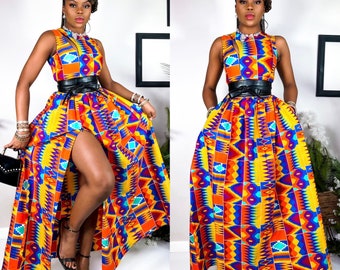 May include: A woman is wearing a long, colorful maxi dress with a high slit. The dress has a geometric pattern in shades of yellow, orange, blue, and red. She is wearing a black belt and has her hair pulled back in a bun.