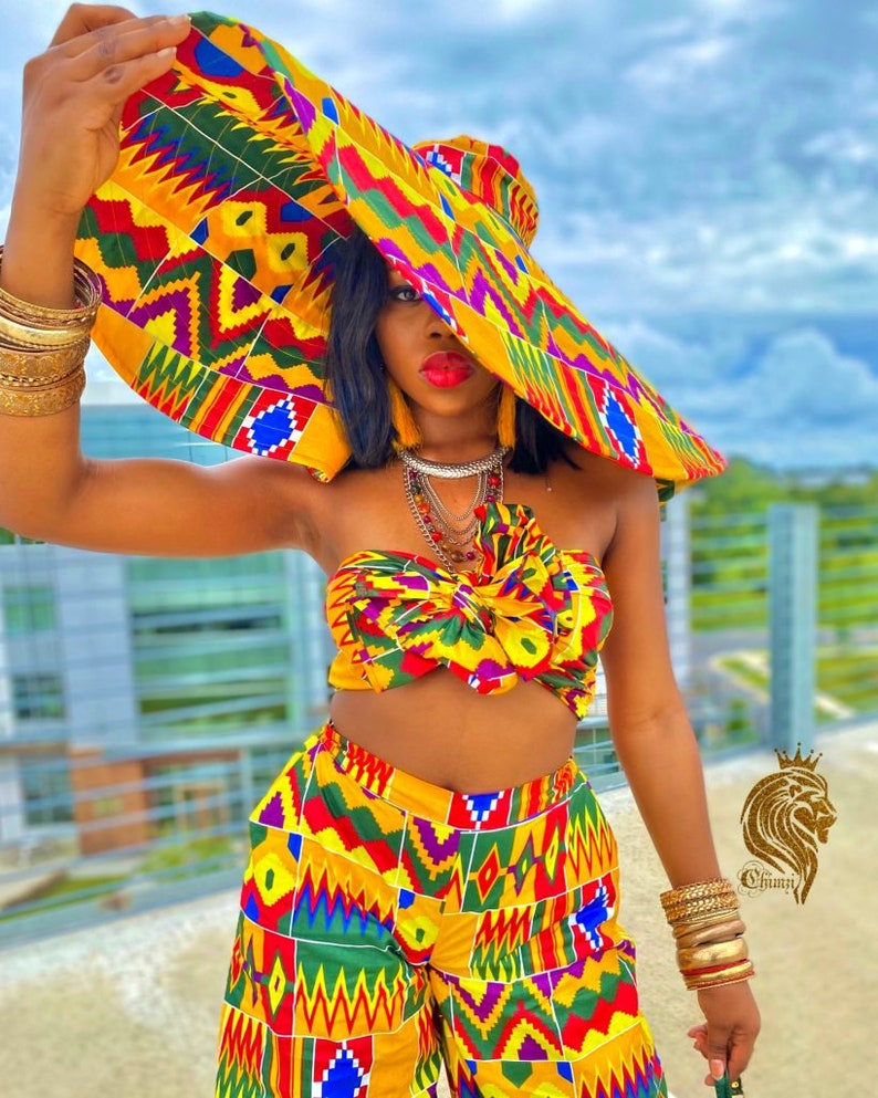 Op de afbeelding: Een vrouw draagt een outfit met een geel, rood, groen en blauw patroon met een bijpassende hoed met brede rand. De outfit bestaat uit een crop top en een wijde broek. Ze draagt meerdere gouden armbanden aan beide polsen.