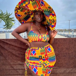 Op de afbeelding: Een vrouw draagt een geel, rood, groen en blauw geprint bikini-topje en een bijpassende rok. Ze draagt ook een grote, breedgerande hoed met hetzelfde patroon. De hoed bedekt haar hoofd en het grootste deel van haar gezicht.