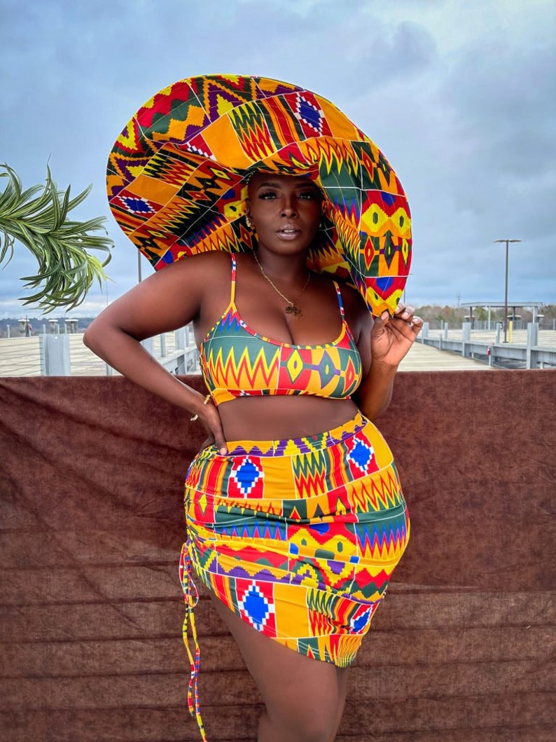 Op de afbeelding: Een vrouw draagt een geel, rood, groen en blauw geprint bikini-topje en een bijpassende rok. Ze draagt ook een grote, breedgerande hoed met hetzelfde patroon. De hoed bedekt haar hoofd en het grootste deel van haar gezicht.