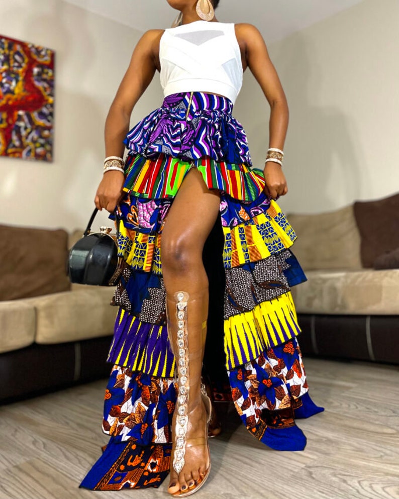 May include: A long, multi-colored skirt with a high slit. The skirt is made of several layers of fabric with different patterns, including stripes, floral designs, and geometric shapes. The skirt is worn with a white tank top and clear heeled sandals.
