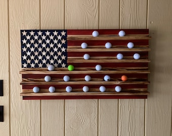 May include: A wooden American flag golf ball display with 25 golf balls in rows. The flag is painted red, white, and blue with a distressed wood finish.