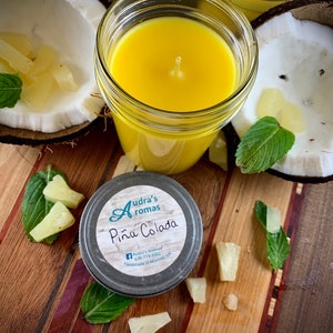 Pode incluir: Uma vela de lata prateada com uma vela amarela dentro. A vela é etiquetada como "Audra's Aromas Piña Colada" e tem um ícone do Facebook e um número de telefone. A vela é cercada por pedaços de abacaxi e uma folha verde.