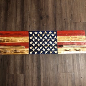 May include: A wooden wall-mounted coat rack painted with an American flag design. The rack has three hooks and is made of reclaimed wood.
