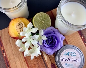 Peut inclure: Deux pots en verre transparent avec des bougies blanches, une pêche, un citron vert, des fleurs blanches, une fleur violette et une boîte en étain argentée avec le texte "Audra's Aromas White Linen" sur une surface en bois.