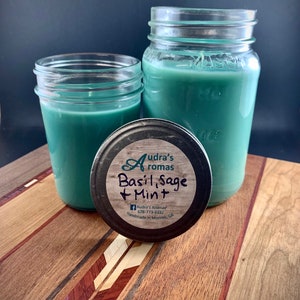 Puede incluir: Dos tarros de vidrio con velas de color verde azulado. El tarro más pequeño está en primer plano y el más grande está en el fondo. La tapa del tarro más pequeño está sobre la mesa delante de los tarros. La tapa tiene una etiqueta negra con texto blanco que dice "Audra's Aromas, Basil, Sage + Mint, 678-773-3332, Handmade in Monroe, GA". Los tarros están sobre una mesa de madera con una incrustación decorativa.