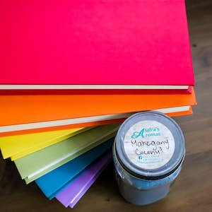 Op de afbeelding: Een stapel kleurrijke boeken met een glazen pot met kaars op een houten oppervlak. De kaars pot heeft een label met de tekst "Audra's Aromas Mahogany Coconut".