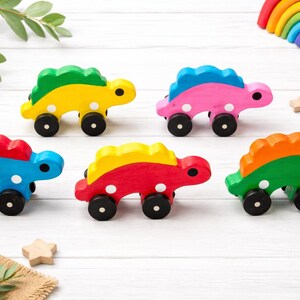 May include: Six colorful wooden dinosaur toys with black wheels and white polka dots. Each dinosaur is painted in a different color combination, including yellow, pink, blue, red, and green. A wooden rainbow and stars are in the background.