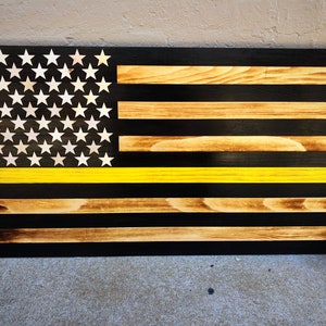 May include: Wooden American flag with a thin yellow line, featuring white stars on a black background. The stripes alternate between black and natural wood tones. The flag is rectangular and appears to be handmade.