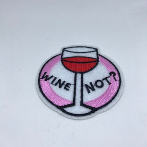 Pode incluir: Etiqueta bordada para passar a ferro com um círculo rosa e o texto "Wine Not?" em preto. Uma taça de vinho tinto está no centro do círculo.