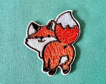 Op de afbeelding: Een geborduurde patch van een vos met zijn staart opgerold. De vos kijkt weg van de kijker en heeft een witte buik en een bruine neus.
