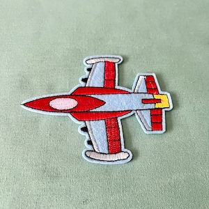 Op de afbeelding: Een geborduurde jetvliegtuigpatch in rood en blauw. Het vliegtuig heeft een wit lichaam met rode vleugels en een gele staart.