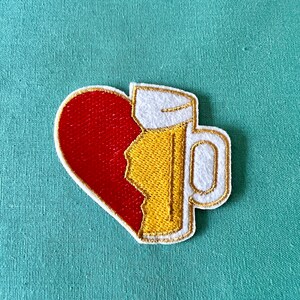 May include: A red heart with a yellow beer mug stitched on top. The heart is broken and the mug is filling the broken space. The mug is white with a yellow beer inside and a white handle.
