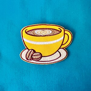 Può includere: Toppa da stiro ricamata gialla di una tazza di caffè con un design a forma di cuore nel caffè e due chicchi di caffè su un piattino.