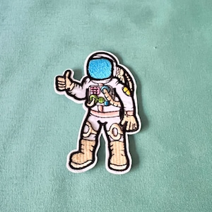 May include: An embroidered iron-on patch of a cartoon astronaut giving a thumbs up. The astronaut is wearing a white spacesuit with a blue visor and colourful details.