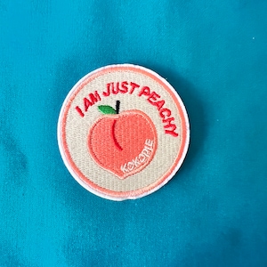 May include: A white, round embroidered patch with the text "I am just peachy" in red. A peach with a red stem and leaf is embroidered in the center of the patch.