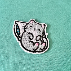 May include: Embroidered iron-on patch featuring a grey cat curled up asleep on a white pillow with blue trim.