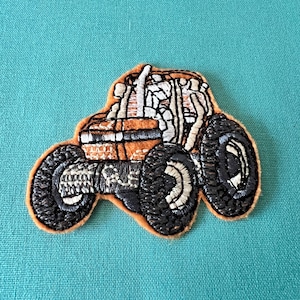 Op de afbeelding: Gestikt patch van een bruine tractor met zwarte banden. De tractor heeft een witte cabine en is versierd met stiksels.