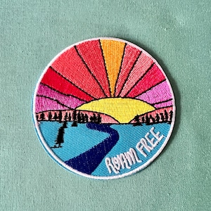 May include: A round embroidered patch with a sunset scene and a river winding through the foreground. The text "ROAM FREE" is written in a white cursive font around the bottom of the patch.
