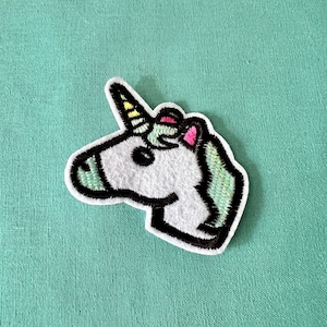 May include: A white unicorn head embroidered patch with a yellow and pink horn. The patch has a black outline and is on a green fabric background.