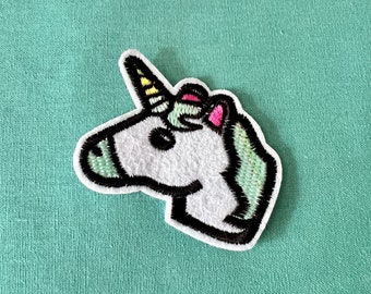 May include: A white unicorn head embroidered patch with a yellow and pink horn. The patch has a black outline and is on a green fabric background.
