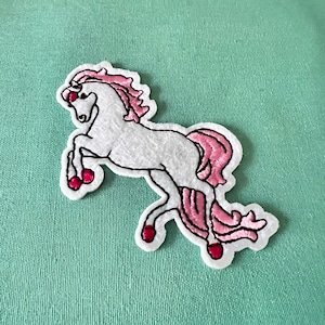 May include: White felt unicorn applique with pink mane and tail. The unicorn has red hooves and is standing on its hind legs.