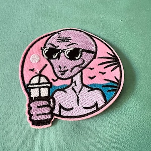 Könnte beinhalten: Ein gestickter Aufnäher mit einem Außerirdischen, der eine Sonnenbrille trägt und ein Getränk hält. Der Außerirdische befindet sich an einem Strand mit Palmen und Vögeln im Hintergrund. Der Aufnäher ist rosa und blau.