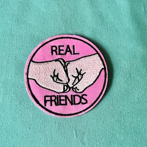Peut inclure: Patch thermocollant rond rose avec broderie noire de deux poings se cognant et le texte "REAL FRIENDS".