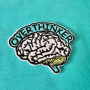 Pode incluir: Um remendo bordado branco e preto com um design de cérebro e o texto "Overthinker" em um contorno azul.