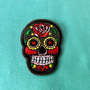 May include: Embroidered black sugar skull patch with colorful details, including a red rose, yellow and red eyes, and white teeth.