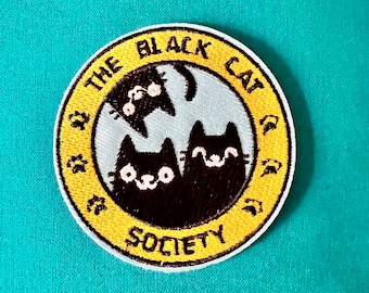 Può includere: Una toppa ricamata gialla e nera con il testo "The Black Cat Society" e tre gatti neri all'interno di un cerchio.