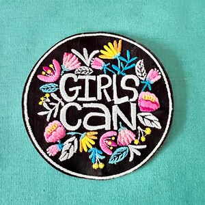 May include: A black embroidered patch with a white floral design and the text "Girls Can" in white.