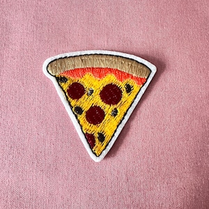 May include: Embroidered pizza slice patch with a white border. The pizza features a yellow crust, red sauce, brown pepperoni, and a golden-brown crust. The patch is on a pink fabric background.