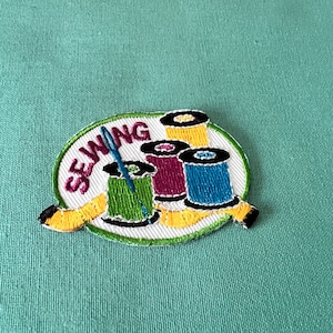 Könnte beinhalten: Gestickter Aufnäher mit dem Wort "SEWING" in Rosa, umgeben von Garnrollen in Grün, Lila, Gelb und Blau. Ein gelbes Maßband ist um die Spulen gewickelt. Eine blaue Nadel befindet sich in der grünen Spule.