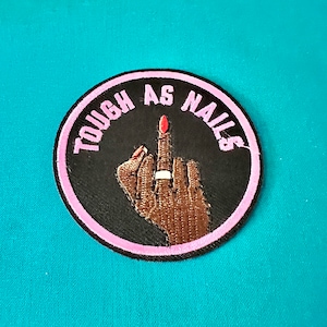 May include: A black and pink embroidered patch with the text "TOUGH AS NAILS" in pink lettering. A raised middle finger with a pink nail is in the centre of the patch.