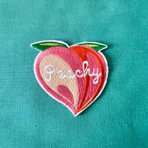 Peut inclure: Patch brodé en forme de pêche avec le mot "Peachy" en écriture cursive blanche. Le patch est rose, orange et vert.