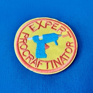 Pode incluir: Um remendo bordado amarelo e laranja com o texto "Expert Procrastinator" em um círculo. Uma pistola de cola quente azul e branca de desenho animado está no centro do círculo.