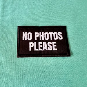 Può includere: Un cerotto di stoffa nero con testo bianco che dice "NO PHOTOS PLEASE" su uno sfondo blu chiaro.