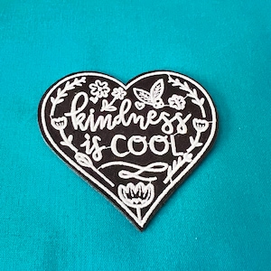 May include: Black heart-shaped patch with white embroidery that reads "kindness is cool". The patch is decorated with small white flowers and leaves.